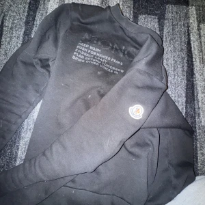 Svart långärmad tröja från Moncler - Säljer en svart långärmad tröja från Moncler med text på bröstet. Tröjan har en hög krage och Moncler-logga på ärmen. Perfekt för kyligare dagar.