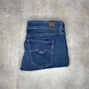 Blå jeans från Replay - Snygga blå dam jeans från Replay med klassisk femficksdesign och knappgylf. Perfekta för en snygg stil med en modern touch. Märkesdetaljer synliga på fickan och insidan av linningen.