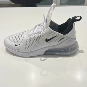 Nike Air Max 270  - Snygga Nike Air Max 270 sneakers i vit med svarta detaljer. Skorna har en stor synlig Air-enhet i hälen för extra komfort och stil. Ovandelen är i ventilerande meshmaterial med snörning för en säker passform.
