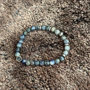 Blått pärlarmband - Snyggt armband med blå och grå pärlor. Armbandet har en elastisk tråd för enkel passform och en liten metallbricka med en ingraverad symbol. Perfekt för att ge en stilren touch till din outfit.