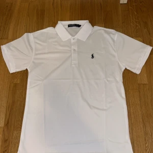 Ralph Lauren pikétröja - PRISET ÄR INTE HUGGET I STEN!!!                 Säljer en klassisk vit pikétröja från Ralph Lauren med korta ärmar och broderad logga på bröstet. Perfekt för en stilren och avslappnad look. Tröjan har en krage och knappar framtill.