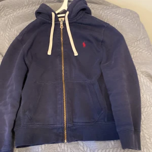 Mörkblå hoodie från Polo Ralph Lauren - Säljer en snygg mörkblå hoodie från Polo Ralph Lauren med dragkedja och röd logga på bröstet. Tröjan har en justerbar huva med vita snören och är tillverkad i en mjuk bomullsblandning. Perfekt för en avslappnad stil. Storleken är M men väldigt liten i storleken så passar S. Hör av er vi funderingar!