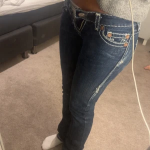 Mörkblå jeans från True Religion - Snygga mörkblå lågmidjade jeans från True Religion med kontrasterande vita sömmar och klassisk femficksdesign. Straight passform. Knappen är tejpad tyvärr men de funkar att knäppa ändå! Små i storleken, 16 rakt över med måttband innerbenslängd ca 30, kan gå ner i pris