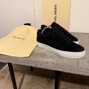 Svarta sneakers från Axel Arigato - Snygga svarta sneakers i mocka från Axel Arigato med vit sula. Skorna har en stilren design med snörning och diskret logga på sidan. Perfekta för en trendig look. Nypris 2399kr