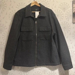 Grå overshirt med dragkedja - Snygg grå overshirt med dragkedja och två stora fickor framtill. Perfekt för lager-på-lager under höst och vår. Klassisk krage och långa ärmar ger en stilren look.