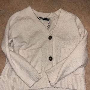 Vit kofta från Vero Moda - Mysig vit kofta från Vero Moda med två stora knappar framtill och praktiska fickor. Perfekt för kyliga dagar när du vill hålla dig varm och stilren. Passar bra till både jeans och kjol.