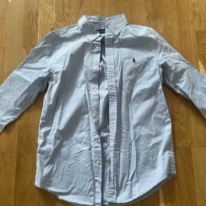 Ljusblå skjorta från Ralph Lauren - Säljer en stilren ljusblå skjorta från Ralph Lauren med klassisk krage och knappar framtill. Skjortan har en broderad logotyp på bröstet och är tillverkad i ett mjukt bomullsmaterial. Perfekt för en elegant look.