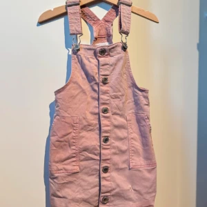 Rosa jeansklänning med knappar - Säljer en söt rosa jeansklänning med knappar framtill och justerbara axelband. Klänningen har praktiska fickor och en avslappnad passform. Perfekt för en avslappnad stil. Fläckar och lösa trådar finns, har ej testat ta bort fläckarna. 