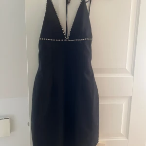 Svart klänning från Adoore - Elegant svart klänning från Adoore med pärldetaljer längs kanterna och en djup v-ringning. Klänningen har en tight passform och är axelbandslös, perfekt för en festlig kväll.