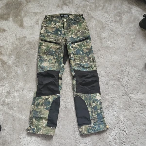 Camouflage byxor från Jägarliv - Säljer ett par coola camouflagebyxor från Jägarliv, perfekta för utomhusaktiviteter. Byxorna har dragkedjor och förstärkta knäpartier för extra hållbarhet. De är designade för att klara tuffa förhållanden och har en praktisk passform. Kan gå ner lite i pris.