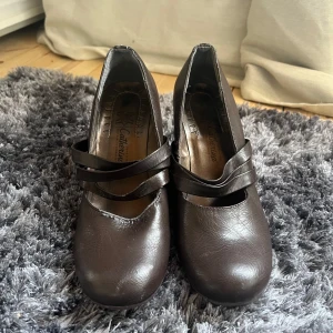 Bruna kilklackskor från Catherina - Snygga bruna kilklackskor från Catherina med elegant design. Skorna har en bekväm kilklack och stiliga remmar över foten. Perfekta för att ge en touch av elegans till din outfit.