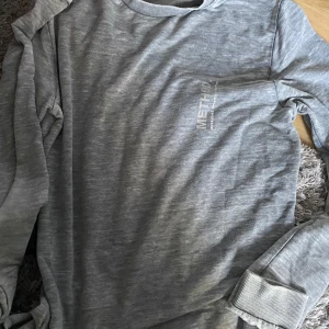 Grå långärmad tröja från Jack & Jones - Säljer en stilren grå långärmad tröja från Jack & Jones Core-kollektion. Tröjan har en rund halsringning och en diskret logga på bröstet. Perfekt för en avslappnad look.