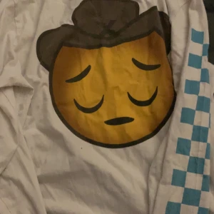 Vit jacka med emoji-tryck - Cool vit jacka med ett stort emoji-tryck på ryggen och blå rutmönster på ärmarna. Designad av Lil Nas X med texten 'The Sad Cowboy' tryckt på framsidan. Perfekt för dig som vill sticka ut med en unik stil.