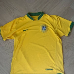 Vintage brasilien fotbolltröjä 2006 - Säljer en gul Brasilien fotbollströja från Nike med gröna detaljer. Tröjan har korta ärmar och ett broderat emblem på bröstet. Perfekt för fotbollsfans. 2006 vintage 