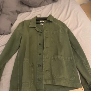 Grön overshirt från Uniqlo - Säljer en stilren grön overshirt från Uniqlo. Jackan har en klassisk krage och knappar framtill. Den har tre praktiska fickor och är perfekt för lager-på-lager-stil. Passar bra till både vardag och mer avslappnade tillfällen.