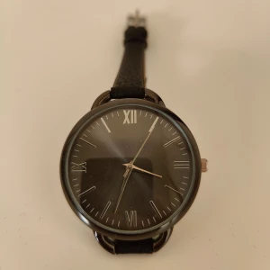Svart klocka med romerska siffror - Elegant klocka med svart urtavla och romerska siffror. Den har ett svart läderarmband och en silverfärgad boett. Perfekt som en stilren accessoar.