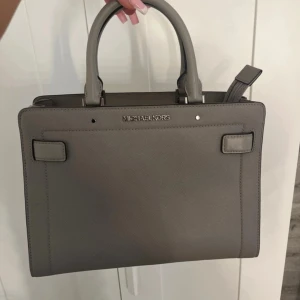 Grå handväska från Michael Kors - Snygg grå handväska från Michael Kors i skinn. Väskan har dubbla handtag och en avtagbar axelrem för flexibilitet. Insidan är fodrad med logotypmönster och har flera fack med dragkedja för enkel organisering. Perfekt för att bära dina dagliga nödvändigheter med stil. Kan gå ner i pris💕