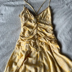 Gul mönstrad klänning med smala axelband - Säljer en somrig gul klänning med  mönster och smala axelband. Klänningen har en v-ringad design och snäckor framtill. Storlek S men skulle tippa på mer XS