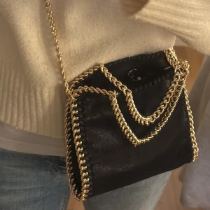 Svart axelväska med guldkedja - Aldrig använd. Liknar Stella McCartney. Snygg svart axelväska med guldkedja som detalj. Väskan har en elegant design med kedjor som ger en lyxig känsla. Perfekt för att lyfta vilken outfit som helst. 