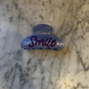 Blå hårklämma med lila text - Säljer en blå hårklämma med texten 'Smile' i lila dekorerad med glittriga stenar. Perfekt för att ge en färgglad touch till din frisyr. Oanvänd!