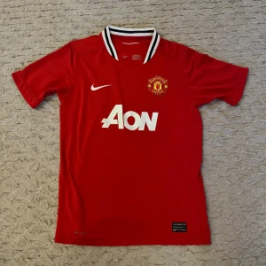 Röd Manchester United fotbollströja - Säljer en röd Manchester United fotbollströja från Nike med vit Aon-logga på framsidan. Tröjan har korta ärmar och en vit och svart randig krage. Perfekt för alla United-fans! 🏆