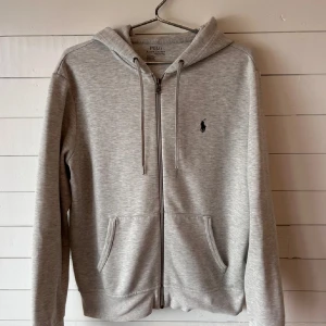 Grå hoodie från Polo Ralph Lauren - Säljer en stilren grå hoodie från Polo Ralph Lauren med dragkedja och klassisk logga på bröstet. Tröjan har en bekväm passform med långa ärmar och praktiska fickor framtill. Perfekt för en avslappnad look.