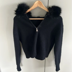 Svart tröja med fuskpälsdetaljer - Snygg svart tröja med dragkedja och fuskpälsdetaljer runt kragen. Perfekt för att hålla sig varm och stilren under kyliga dagar. Tröjan har långa ärmar och en ribbad nederkant. Den är från Majive, och är använd 1 gång. Man kan även ta bort pälsen 