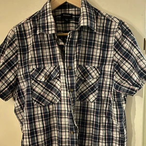 Rutig kortärmad skjorta från Twister - Säljer en snygg rutig skjorta från Tuxer i blått, vitt och rött (rutigt mönster). Skjortan har korta ärmar och knappar framtill samt två bröstfickor med knappstängning. Perfekt för en avslappnad stil till sommaren och släktkalas! 