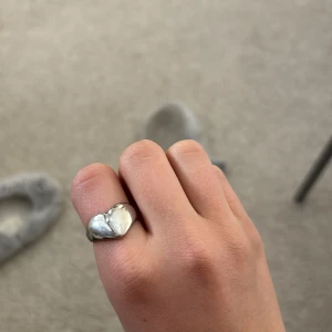 Silverfärgad ring med hjärtdetalj - En stilren och justerbar ring i silverfärg med en söt hjärtdetalj framtill. Perfekt för att ge en romantisk touch till din outfit.