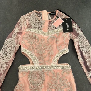 Rosa spetsklänning från PrettyLittleThing - Elegant rosa klänning med intrikata spetsdetaljer och pärlbroderier. Klänningen har långärmade spetsärmar och en figurnära passform. Perfekt för speciella tillfällen med sin vackra design och detaljerade mönster.