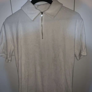 Vit pikétröja från Selected Homme - Säljer en stilren vit pikétröja från Selected Homme. Tröjan har en dragkedja framtill och korta ärmar, perfekt för en avslappnad look. Den har en klassisk krage och är tillverkad i frote.