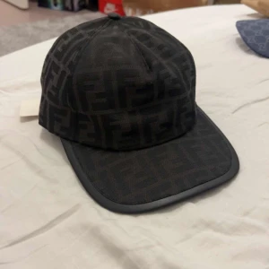 Svart Fendi keps med monogram - Snygg svart keps från Fendi med det klassiska FF-monogrammet över hela kepsen. Kepsen har böjd skärm och stilren design som ger en lyxig vibe. Perfekt för dig som vill ha något exklusivt och trendigt i garderoben. Och helt ny