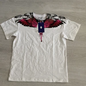 Marcelo Burlon tshirt - (Passar även folk som har storlek S) Snygg vit ärmlös t-shirt från med ikoniskt tryck av rosa och svarta vingar över axlar och bröst. T-shirten har rund halsringning och är tillverkad i mjuk bomull. Perfekt för dig som vill sticka ut med en cool och unik design.