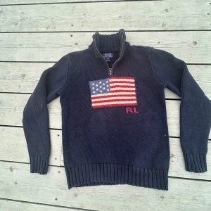 Mörkblå stickad tröja Polo Ralph Lauren - Snygg mörkblå stickad tröja från Polo Ralph Lauren med amerikansk flagga framtill och RL-broderi. Tröjan har ribbad krage, muddar och nederkant samt en half zip-dragkedja vid halsen. Perfekt för dig som gillar klassisk stil med en twist.