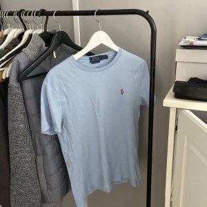 Ljusblå t-shirt från Polo Ralph Lauren - Snygg ljusblå t-shirt från Polo Ralph Lauren med klassisk rund hals och korta ärmar. Ikoniska broderade loggan på bröstet ger en clean och stilren vibe. Perfekt för dig som gillar enkel och tidlös design.