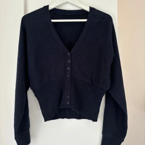 Mörkblå stickad kofta med knappar - Snygg mörkblå stickad kofta med v-ringning och knappar framtill. Modellen är croppad med ribbad nederkant och muddar vid ärmslut. Perfekt att slänga över en t-shirt eller linne för en chill look.