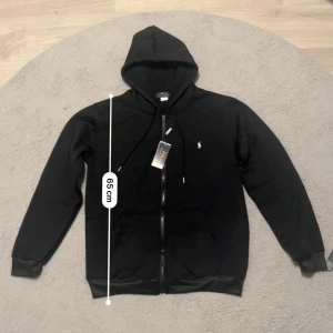 Svart hoodie från Polo Ralph Lauren - Svart hoodie från Polo Ralph Lauren med klassisk vit logga på bröstet. Tröjan har huva med snörning, dragkedja framtill och två fickor. Tillverkad i mjuk bomull, perfekt för chill dagar. Långärmad och med ribbade muddar för extra komfort.Den är perfekt för vintern och hösten.höjden är 65 cm och Breden är 50 cm.💬Skriv gärna om du har frågor! 📦 Skickas inom 24 timmar! 📉 Kan gå ner lite i pris vid snabb affär! 🕒 Aktiv nästan hela tiden svarar snabbt! 