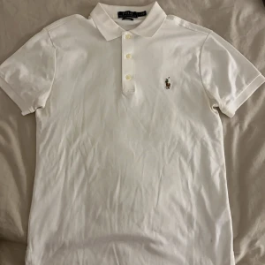Vit slim fit piké från Polo Ralph Lauren - Vit pikétröja från Polo Ralph Lauren i slim fit-modell. Klassisk krage, tre knappar framtill och broderad logga med polospelare på bröstet. Tillverkad i mjuk bomull, perfekt för en clean och stilren look.