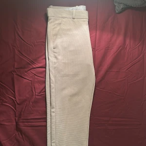 Beige kostymbyxor med struktur - Snygga beige kostymbyxor med diskret strukturerat tyg och klassisk passform. Byxorna har hög midja, bälteshällor och sidofickor. Perfekta för dig som gillar stilrena och cleana vibes. Stängs med knapp och dragkedja framtill.