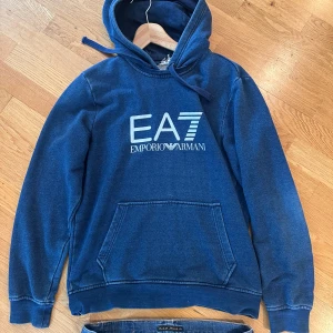 Blå EA7 Emporio Armani hoodie - Säljer en snygg marinblå hoodie från EA7 Emporio Armani med stort vitt tryck på bröstet. Tröjan har huva med snörning, känguruficka och långa ärmar. Perfekt för dig som gillar streetwear och vill ha en stilren look.