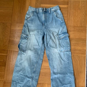Blå cargopants i jeansmaterial - Säljer ett par ljusblå cargopants i jeansmaterial med bred passform och stora fickor på sidorna. Byxorna har klassisk femficksdesign, bälteshällor och raka ben. Perfekta för dig som gillar streetwear och vill ha gott om plats för prylar.