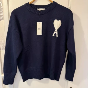 Marinblå stickad tröja från Ami - Säljer en marinblå stickad tröja från Ami med rund hals och ribbade muddar. På bröstet finns ett vitt hjärta med bokstaven A, som är typiskt för märket. Tröjan har lång ärm och en clean, oversized passform som är riktigt chill.