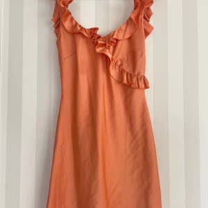 Persikofärgad kort klänning med volang - Snygg persikofärgad klänning med volangdetaljer längs halsringningen och axlarna. Klänningen är ärmlös och har en feminin, lätt A-linjeformad siluett. Perfekt för sommarens alla tillfällen och ger en fräsch och ungdomlig vibe.