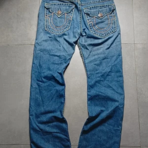 True Religion Jeans - Säljer ett par blå jeans från True Religion med snygga slitningar framtill och broderade bakfickor med knappar. Jeansen har klassisk femficksdesign, kontrastsömmar och raka ben. Perfekt för dig som gillar en cool och avslappnad stil.
