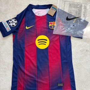 FC Barcelona Lamine Yamal tröja Nike M - Snygg FC Barcelona matchtröja med Lamine Yamal och nummer 10 på ryggen. Tröjan är kortärmad, har klassiska blå och röda ränder, gula detaljer och klubbmärke på bröstet. Tillverkad i lätt polyester med Champions League och Foundation-patch på ärmen.