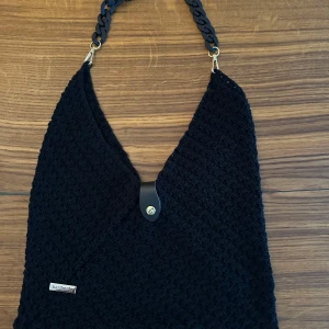 Svart stickad axelväska handmade - Snygg svart stickad axelväska med grovt mönster och cool kedja som axelrem. Väskan har en enkel knappstängning i läderimitation och en unik form som sticker ut. Perfekt för dig som gillar handgjorda accessoarer med personlig touch.