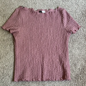 Rosa ribbad topp från H&M Divided - Säljer en ljusrosa ribbad topp från H&M Divided i storlek S. Toppen har korta ärmar, stretchigt material och vågiga kanter vid halsringning, ärmslut och nederkant. Perfekt att styla med jeans eller kjol för en chill look.