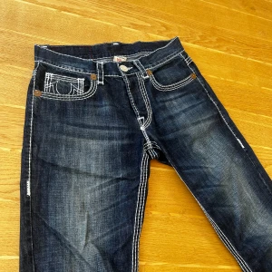 True Religion mörkblå jeans med kontrastsömmar - Säljer ett par mörkblå jeans från True Religion med tydliga vita kontrastsömmar och ikoniska broderade fickor både fram och bak. Jeansen har rak passform och klassisk femficksdesign, tillverkade i slitstarkt denim. Perfekt för dig som gillar street och detaljer. Storlek 34, kan skicka mått om så önskas