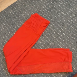 Röda sportleggings från H&M - Snygga röda/oranga  sportleggings från H&M med meshdetaljer längs benen och perforerat parti vid midjan. Stretchigt syntetmaterial som sitter tight och är perfekt för träning. Modellen är högmidjad och har en modern, atletisk look.