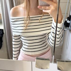 Randig offshoulder ribbad topp - Säljer en vit och svart randig offshoulder topp med långa ärmar. Toppen är ribbad och har en croppad passform som sitter snyggt på kroppen. Perfekt för dig som gillar en stilren och trendig look.